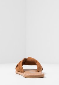 Braune Wildleder-Slipper mit verdrehten Oberriemen, flacher Sohle und hellbraunem Fußbett. Minimalistisches Design, geeignet für die Freizeit.
