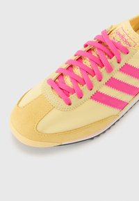 Gul atletisk sko med rosa detaljer og snøringer. Den har semsket lær og mesh-materialer, tre rosa striper, og en avrundet tådesign.