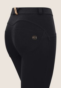 Pantaloni aderenti neri con cuciture dettagliate, passanti per cintura e un piccolo emblema del marchio quadrato dorato nell'area della tasca posteriore.