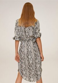 Femme aux longs cheveux portant une robe imprimée serpent, ceinturée et de longueur genou avec des manches retroussées, vue de dos sur un fond uni.