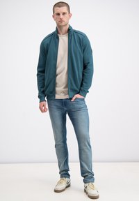 Męska turkusowa kurtka zapinana na zamek z teksturowanymi rękawami, w połączeniu z beżową koszulką, jasnoniebieskimi dżinsami i białymi sneakersami z akcentami.