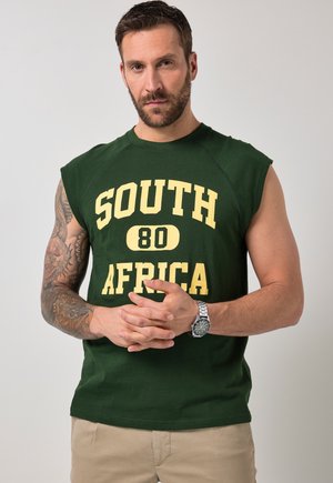 Mann mit Bart und tätowiertem Arm, trägt grünes ärmelloses Shirt mit der Aufschrift "South Africa 80" und beigen Hosen, posiert vor einfachem Hintergrund.