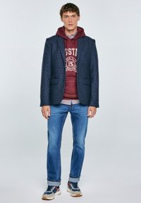 Mladý muž v tmavomodrom károvanom saku cez bordový hoodie, s modrými džínami s manžetami a pestrofarebnými teniskami, stojaci na bielom pozadí.