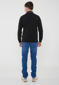 Polo a maniche lunghe nera con dettaglio colletto bianco, abbinato a jeans in denim blu e scarpe beige, vista da dietro.
