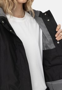 Svart jacka med grå inredning, tryckknappsstängningar och en mjuk textur. Jackan är delvis öppen och visar en vit t-shirt under.