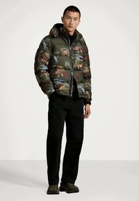 Miesmodeli seisoo päällään oliivinvihreä untuvatakki, jossa on villieläinkuvasto, mustat housut ja tummat lenkkarit yksinkertaista valkoista taustaa vasten.