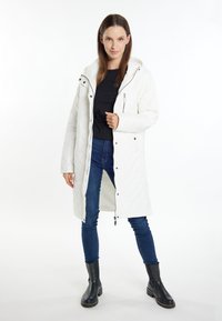 Witte gewatteerde jas met een capuchon, voorzien van een rits en drukknoopjes, gedragen over een zwarte top en blauwe jeans, in combinatie met zwarte enkellaarsjes.