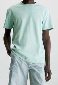 Mátově zelené bavlněné tričko s kulatým výstřihem a krátkým rukávem, s malým bílým logem na hrudi a volným střihem.
