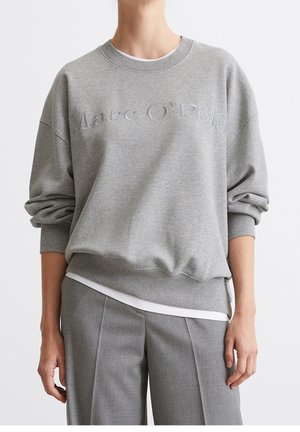 Personne portant un sweat-shirt gris Marc O'Polo par-dessus une chemise blanche, assorti d’un pantalon gris sur mesure, debout devant un fond uni.