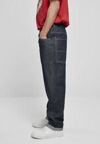 Jeans din denim albastru închis, cu croială lejeră, dotate cu o buză laterală, cusături în contrast și tivuri răsucite, purtate cu adidași albi.