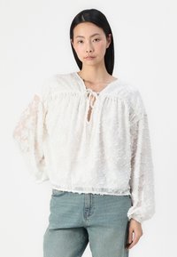 Weiße Spitzenbluse mit floralem Muster, lockerer Schnitt, elastischem Saum und Kordelzug am Ausschnitt. Lange, transparente Ärmel sind texturiert und luftig.