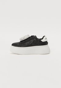 Sorte lærersneaker med hvit ribbet såle, med en blomsterdetalj på tungen og "Karl" preget på siden.