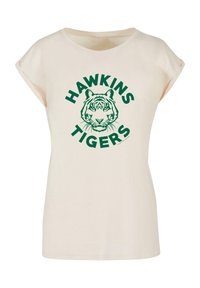 Kremfarget t-skjorte med korte ermer og rund hals, som har et grønt tigermotiv og «HAWKINS TIGERS» tekst i fete grønne bokstaver.