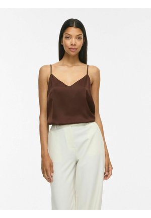 Kvinde står og vender fremad iført brun camisole-top og højtaljede hvide bukser mod en ensfarvet lys baggrund.