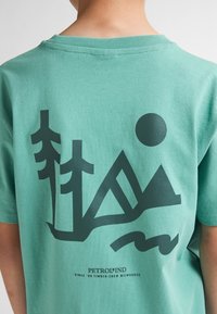 Tricou din bumbac verde, cu un grafic întunecat reprezentând copaci, munți și un soare, având textul "PETROL IND" în partea de jos.