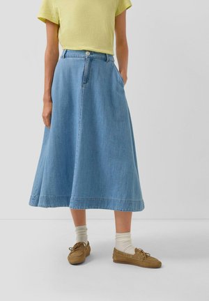 Person trägt ein gelbes T-Shirt, einen hellblauen Midi-Jeansrock mit Taschen, beigefarbene Socken und braune legere Schnürschuhe und steht vor einem weißen Hintergrund.