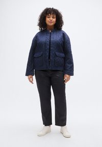 Μπλε σκούρο καπιτονέ μπουφάν με στρογγυλή λαιμόκοψη, κούμπωμα με κουμπιά και δύο μπροστινές τσέπες, συνδυασμένο με μαύρο παντελόνι και λευκά αθλητικά παπούτσια.