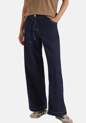 Person trägt dunkelblaue Jeans mit weitem Bein und Kordelzug in der Taille, beiges Oberteil und beige Turnschuhe mit grünen Streifen, steht mit einer Hand in der Tasche.