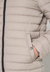 Beige gewatteerde jas met horizontale doorgestikte secties, een zwarte ritssluiting en een zijzak die wordt afgesloten met een drukknoop.