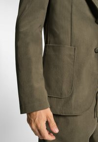 Olijfgroene corduroy blazer met textuurstof, sluiting met één knoop en een paspelzak aan de linkerkant. Getailleerd silhouet met op maat gemaakte mouwen.