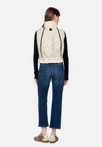 Gepolsterte beige Weste mit strukturiertem Design, schwarzen langen Ärmeln und einem gerafften Saum; kombiniert mit dunklen Jeans und gemusterten Sneakers.