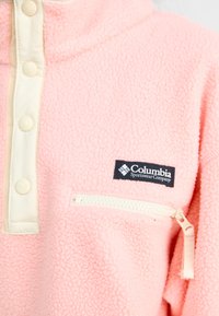 Pull-over en polaire rose clair avec des boutons-pression crème, une poche zippée et une étiquette de marque Columbia sur le côté gauche. Texture douce.