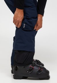 Marineblauwe skibroek met een aansluitende pasvorm en elastische boorden, gecombineerd met zwarte skiskchoenen met rode accenten en een strakke afwerking.