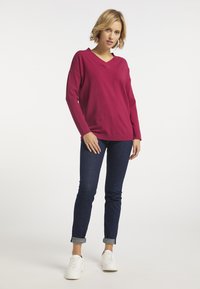 Fuchsiaszínű V nyakú pulóver hosszú ujjal, sötétkék farmernadrággal, amelynek szárát felgöngyölték, és fehér sportcipővel párosítva. Simára szőtt anyag és laza fazon.