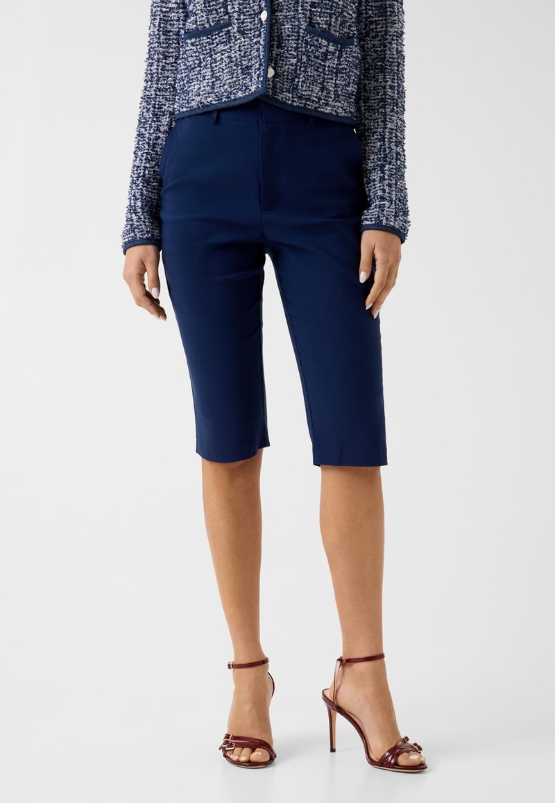Femme portant un short bleu marine taillé sur mesure au genou, une veste texturée bleue et blanche, et des sandales à talons marron, debout devant un fond uni.