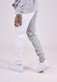 Jeans bicolor con la pierna izquierda en blanco y la pierna derecha en denim gris. Presentan un corte ajustado, un ligero desvanecimiento y se combinan con zapatillas blancas.
