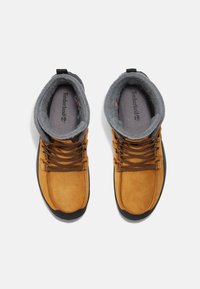 Bottes en suede marron avec un col en polaire grise, semelle en caoutchouc noire et lacets noirs et orange, avec un logo Timberland sur la semelle intérieure.