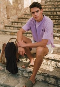 Levandų spalvos trumpomis rankovėmis marškiniai, balti šortai, alyvuogių žalios spalvos šlepetės. Pintas šiaudų krepšys, juodas audinys po šalia. Textūruoti akmens laiptai kaip fonas.