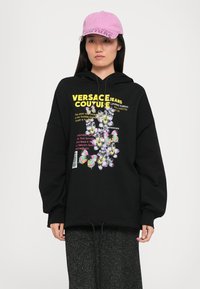 Hoodie oversized preto com um gráfico colorido de flores e borboletas, com texto amarelo. Usado com um boné rosa e calças cinza texturizadas.