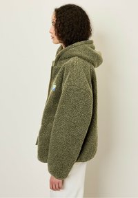 Veste en polaire surdimensionnée de couleur vert olive avec une surface texturée, capuche attachée et poches avant ; présente un petit écusson multicolore sur le côté.