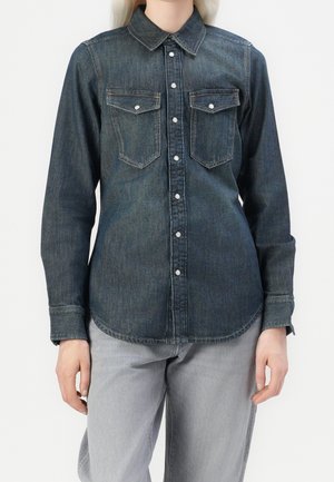 Personne portant une chemise en jean bleu foncé à boutons avec deux poches à rabat à l'avant et un jean gris clair, se tenant devant un fond uni.