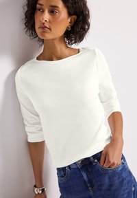 Haut blanc à manches longues avec un col bateau, texture douce et coupe décontractée, associé à un jean en denim bleu. Design simple et élégant.