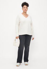 Witte V-hals trui met lange mouwen, gecombineerd met donkere jeans. Het model houdt een kleine witte tas vast en draagt lichte sneakers.