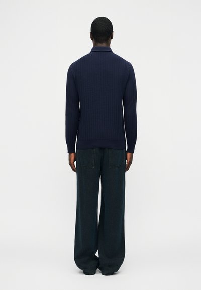 Homme debout, de dos, portant un pull long à manches longues côtelé bleu marine et un jean large foncé, sur un fond blanc uni.