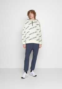 Krémová mikina s čiernym textom "CARLO COLUCCI", v kombinácii s námorníckymi teplákmi. Biele tenisky s čiernymi akcentmi dotvárajú outfit.