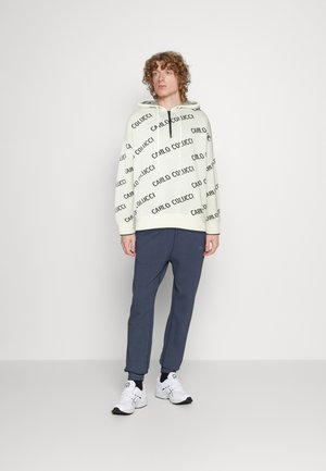 Cremefarbene Kapuzenjacke mit schwarzem "CARLO COLUCCI"-Text, kombiniert mit marineblauen Jogginghosen. Weiße Sneakers mit schwarzen Akzenten runden das Outfit ab.