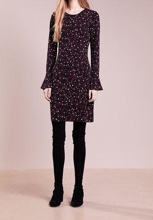 Femme portant une robe noire à manches longues avec un motif d’étoiles multicolores, associée à des bottes noires hautes, debout sur un sol beige.