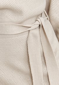 Beige gestricktes Kleidungsstück mit einem texturierten Muster, einem Taillengürtel und glattem Stoff an den Rändern.