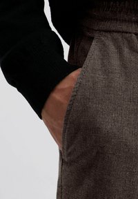 Pantalons en tissu à carreaux marron avec une poche latérale, associés à un pull noir. La texture est douce et la coupe semble décontractée.