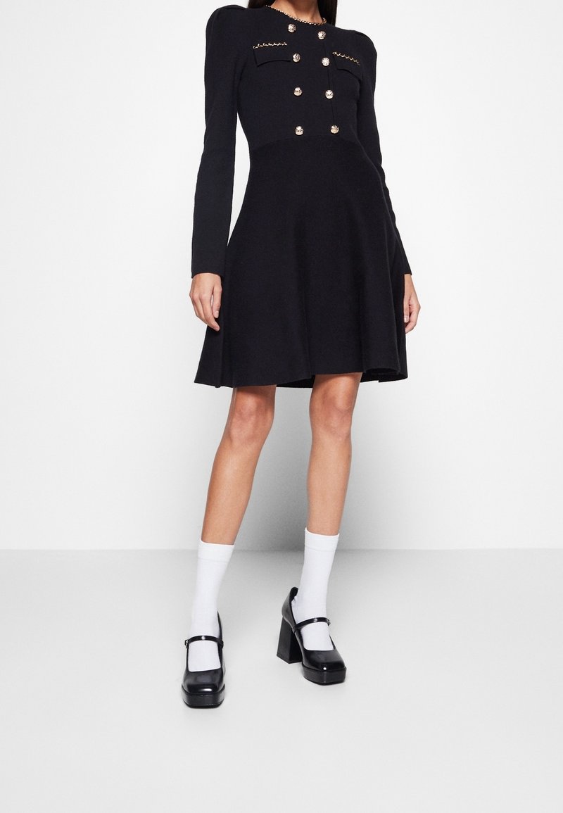 Femme portant une robe noire à manches longues avec des boutons dorés et des poches devant, des chaussettes blanches et des chaussures Mary Jane noires à plateforme.