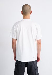 Hvit bomulls t-skjorte med korte ermer og rund hals, med et enkelt design. Kombinert med mørke cargo-shortser og synlige tatoveringer på armen.
