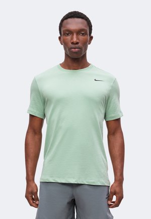 Homme debout, face à l'avant, portant un t-shirt Nike vert clair et un short gris sur un fond clair uni.