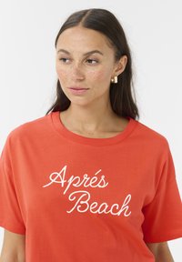 Rød bomulds-T-shirt med hvidt kursiv tekst "Après Beach." Rund halsudskæring med korte ærmer, glat tekstur og afslappet pasform.