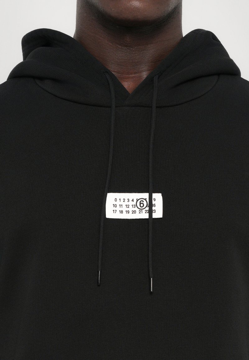 Sudadera negra con textura de canalé, que presenta una etiqueta blanca con números impresos y un acento circular en el pecho. Cordones en la capucha.