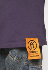 Violet medvilnės marškinėliai su oranžine etikete ir juodu tekstu. Marškiniai derinami su mėlynomis džinsinėmis kelnėmis, pabrėžiančiomis tekstūruotą audinį.