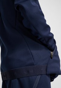 Mörkblå sportjacka med en slät yta, utrustad med en ficka med dragkedja och ett band med varumärkets namn vid midjan.